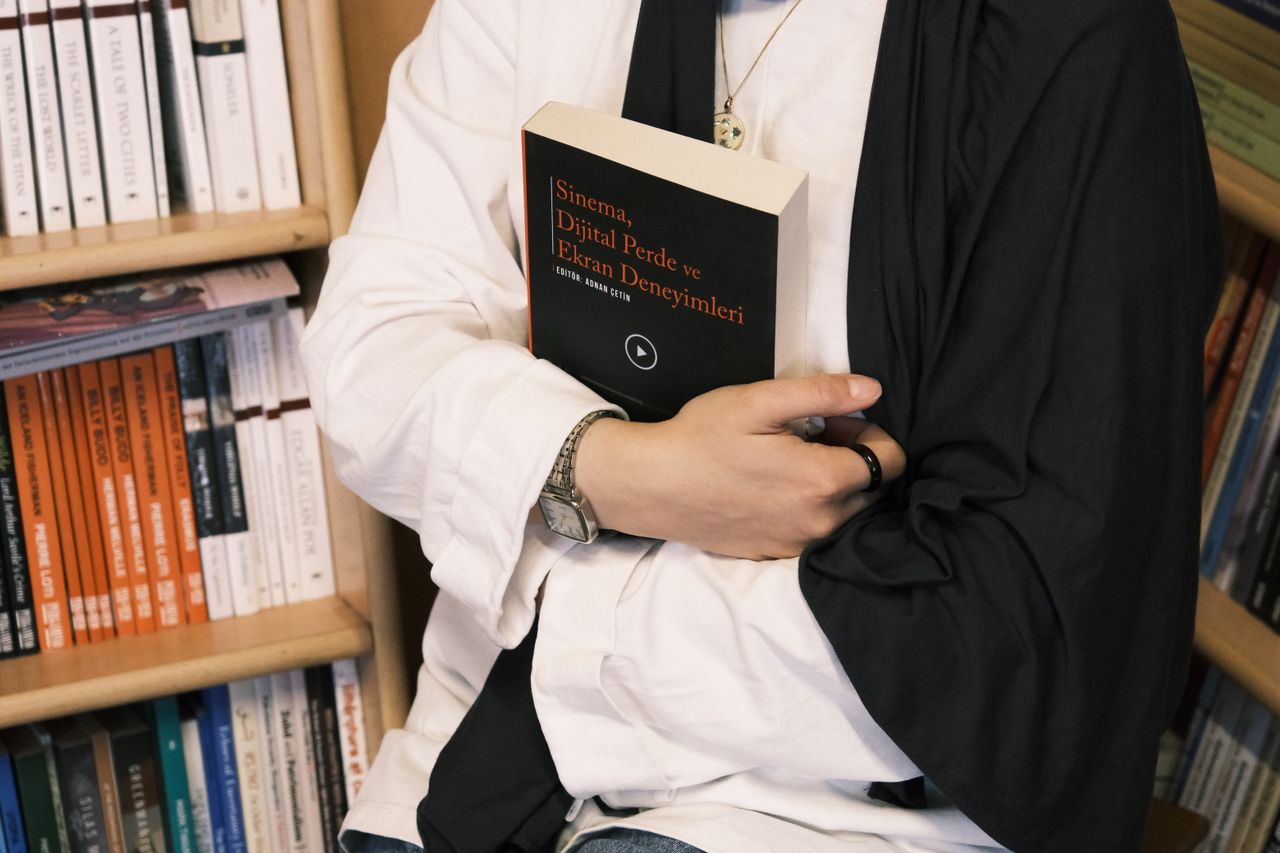 A person holds a book titled 'Sinema, Dijital Perde ve Ekran Deneyimleri' in a cozy library setting.